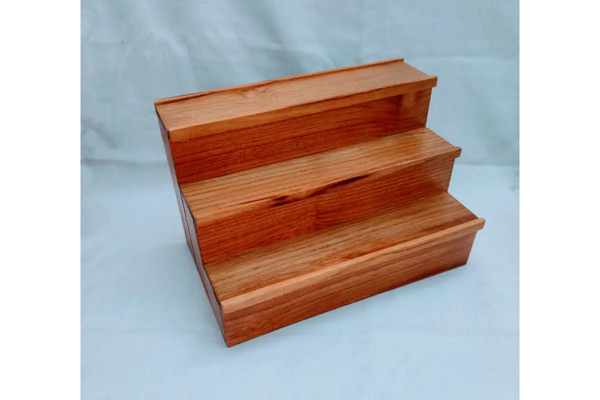 A three-step wooden display riser or shelf made of polished reddish-brown wood, photographed against a light blue-gray backdrop.