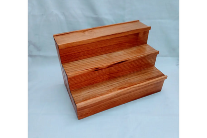 A three-step wooden display riser or shelf made of polished reddish-brown wood, photographed against a light blue-gray backdrop.