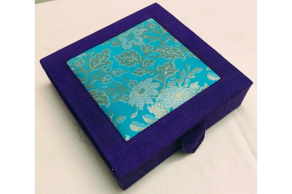 A square purple fabric-covered box with a hinged lid, featuring a turquoise blue brocade patterned panel on the top with gold floral motifs.