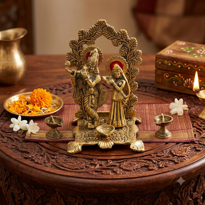 A Lord Krishna and Radha idol displayed on a carved wooden table with traditional decorative items including marigold flowers, white flowers, and oil lamps.