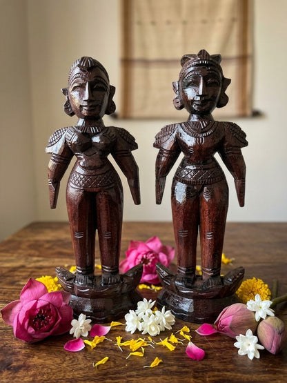 Two wooden statues on a wooden surface with flowers around them.