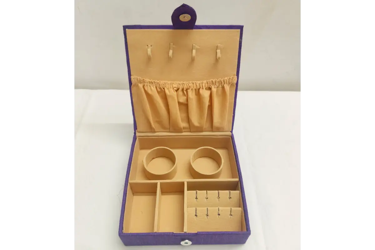 An open jewelry box with a purple exterior and tan interior, featuring compartments, ring rolls, a necklace holder with hooks, and a small top loop, photographed on a white surface against a white background.