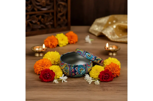 A decorative metal pot with a colorful paisley patterned lid, displayed on a wooden surface surrounded by marigold flowers, lit oil lamps, and a gold fabric cloth.