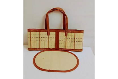 A woven straw tote bag with brown leather handles and trim, displayed next to an oval matching leather pad on a white surface against a plain light background.
