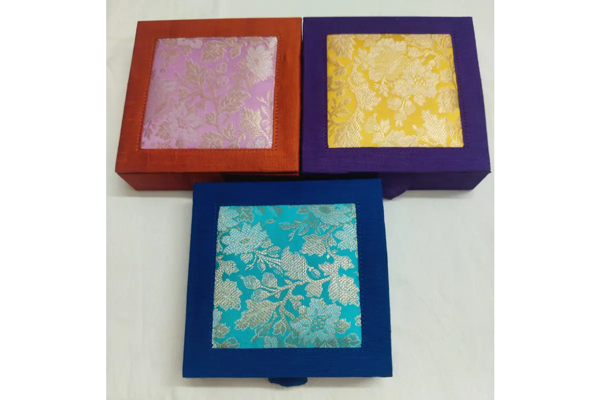 Three square decorative boxes with woven floral brocade fabric lids, shown in red-orange, purple, and blue frames on a white background.
